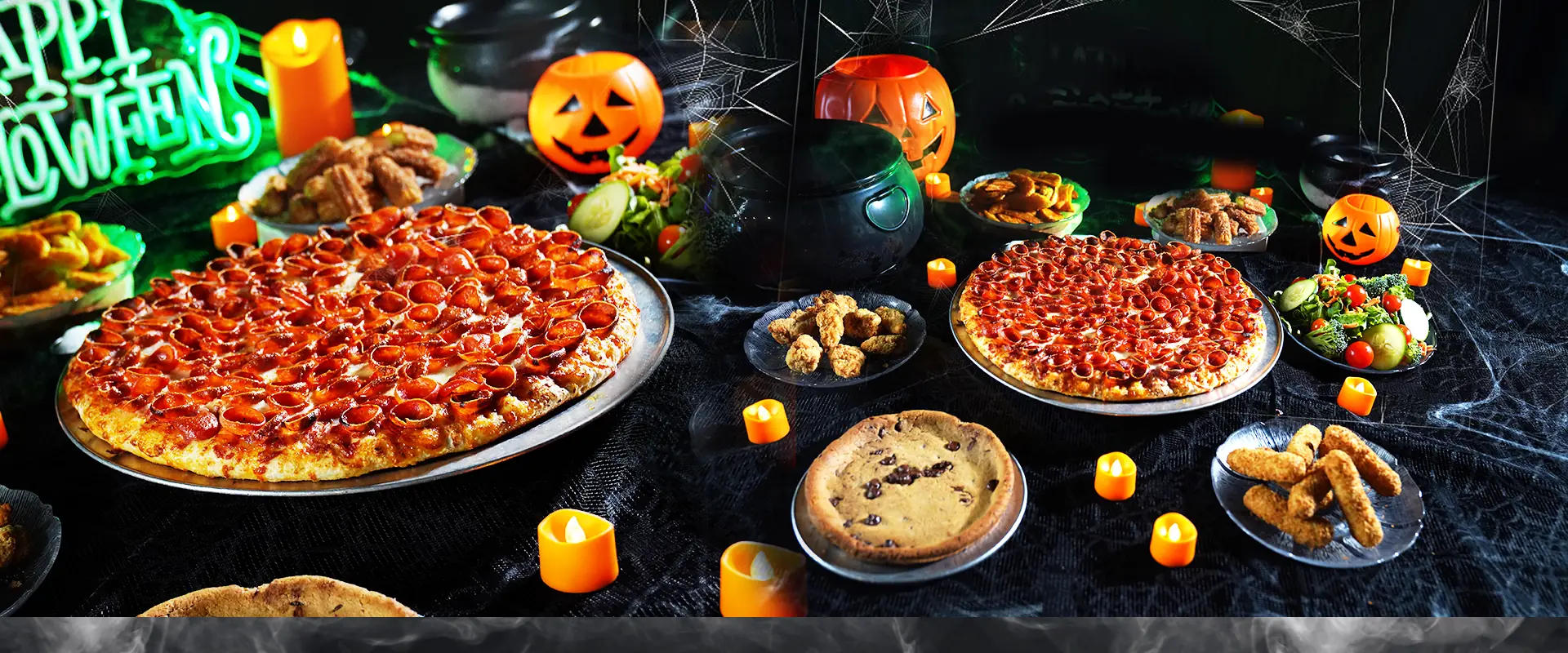 A Halloween feast table: pepperoni pizzas, wings, salads, mozzarella sticks and a pizza cookie among jack-o'-lanterns, candles and a neon Happy Halloween sign